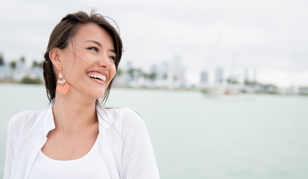 A woman stands smiling wearing white in front of a body of water with a skyline in the background.